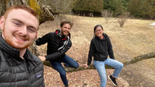 Three students in a photo sitting on a long branch with a field behind them.