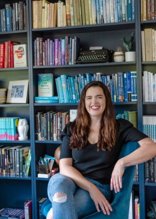 Kate, a white woman with long brown hair sits in a chair in front of a bookcase, full of books, wearing a black shirt and blue jeans with holes in the knees.