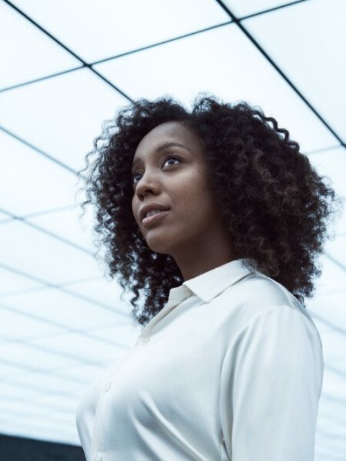 Moo, a black woman with shoulder length black hair poses for a head shot under a bright white square designed ceiling, wearing a white blouse.