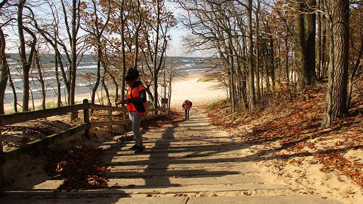 Two students wearing orange safety vests are taking measurements along a path with the beach and Lake Michigan in the distance.